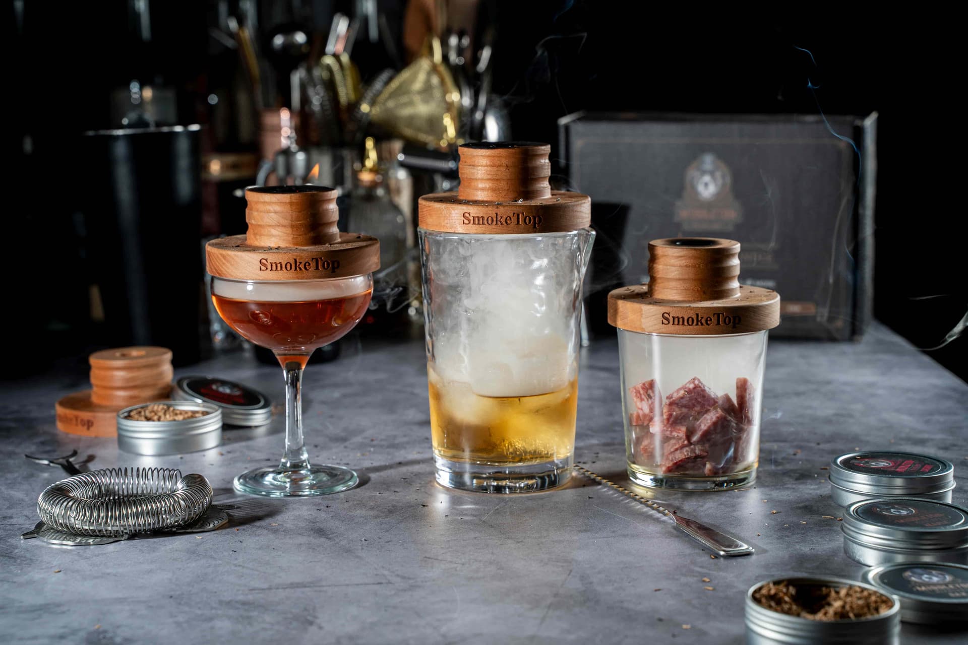 Three SmokeTop cocktail smokers on different glasses with smoked drinks, cheese, and charcuterie on a bar